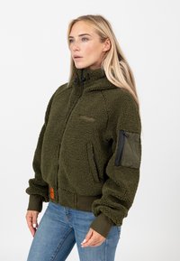 Zielona oliwkowa polarowa kurtka z wysokim kołnierzem, zamkiem błyskawicznym i ściągaczami przy mankietach. Posiada dodatkową kieszeń na zamek błyskawiczny na rękawie.