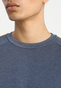 Felpa blu a girocollo con texture a coste, con accenti gialli sulla spalla. Inquadratura ravvicinata dell'area del collo e del petto superiore.