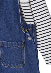 Denim-Latzhose in tiefblau mit orangefarbenen Nähten. An der Seite sind goldene Beschläge und ein weiß gestreiftes Langarmhemd blitzt hervor.