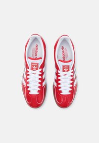 Red synthetic athletic shoes with white laces and three lateral stripes, featuring a padded collar and textured rubber sole for grip.