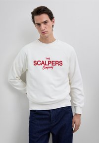 Hvid sweatshirt med røde broderede bogstaver "THE SCALPERS Company," rund hals og ribbede manchetter. Parret med mørke denim jeans.
