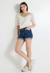 Top crema de manga corta con detalles de encaje, combinado con shorts de mezclilla azul oscuro, con puños enrollados, y zapatillas beige con un diseño texturizado.