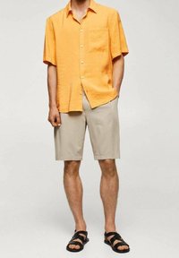 Uomo che indossa una camicia arancione a maniche corte con bottoni, pantaloni corti beige fino al ginocchio e sandali neri, in piedi con le mani parzialmente nelle tasche.