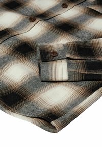 Camicia in flanella a quadri con motivi marroni e neri, dotata di bottoni marroni e polsini piegati. Texture morbida, palette di colori neutri.
