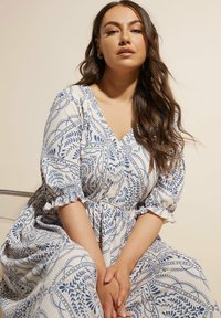 Valkoinen mekko sinisellä kukkakuviolla, V-pääntie, lyhyet puffihihat ja röyhelökoristeet. Pehmeä tekstuuri, virtaava istuvuus ja kerätty vyötäröyksityiskohta.