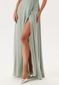 Dlouhé, mátově zelené šaty z hladké tkaniny, které mají vysoký boční rozparek a drapérie v pase, doplněné lesklými stříbrnými podpatky.