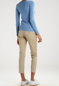 Kvinna iklädd en ljusblå långärmad topp, beige cropped byxor och vita sneakers, står med ryggen mot kameran på vit bakgrund.