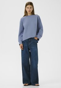 Felpa azzurro chiaro con una cucitura diagonale, abbinata a jeans di denim blu scuro oversize. Tessuto morbido, vestibilità rilassata e gambe ampie.