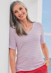 Weißes T-Shirt mit horizontalen roten und blauen Streifen, kurzärmlig und mit Rundhalsausschnitt. Das Modell lehnt an einer Wand und trägt rote Hosen.