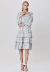 Jeune femme debout, portant une robe à manches longues, à volants, à motif floral allant jusqu'au genou avec des sandales à talons hauts blanches.