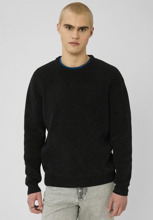 Jeune homme aux cheveux blonds courts portant un pull noir et un jean gris clair sur un fond neutre.