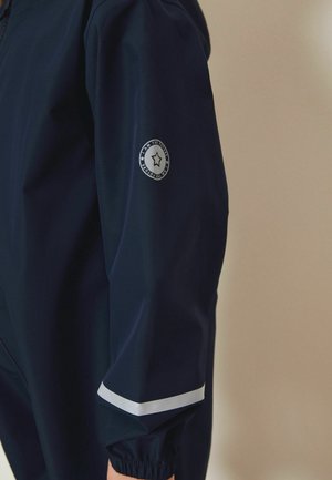 Marineblå jakkeærme med elastisk manchet, reflekterende diagonalt stribe nær håndleddet og cirkulært mærke med stjerne og teksten "JEG ER FREMTIDEN".