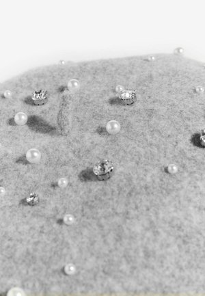 Szara tkanina o różnorodnych teksturach i rozmiarach, ozdobiona rozsypanymi perłami i kryształowymi elementami, leżąca na płaskiej powierzchni.