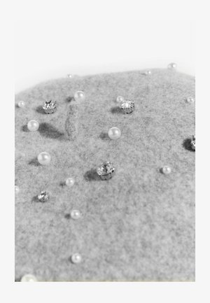 Šedý filcový materiál zdobený rozptýlenými perlami a krystalovými ozdobami, s různými texturami a velikostmi na plochém povrchu.