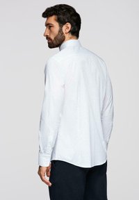 Homme aux cheveux foncés et à la barbe, portant une chemise blanche à manches longues avec de petits points et un pantalon foncé, vu de dos, regardant sur le côté.