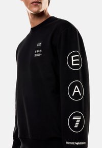 Černá mikina z měkké látky s žebrovaným límcem. Má bílé grafické potisky: "EA," "A" a "7" na rukávu a malou logo na přední straně.