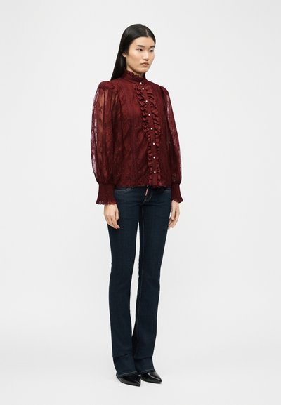 Γυναίκα που φοράει διάφανη μπορντό δαντελένια μπλούζα με βολάν στο μπροστινό μέρος και κουμπιά από πέρλες, σε συνδυασμό με σκούρο μπλε εφαρμοστά τζιν και μαύρα μυτερά παπούτσια.