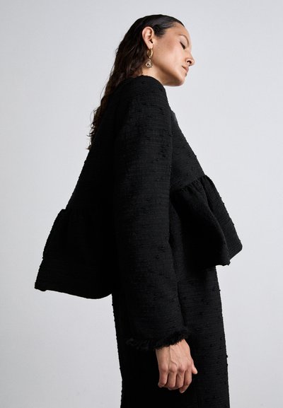 Black textured jacket with a peplum design, featuring a curved hem and loosely stitched details. Long sleeves with frayed cuffs.