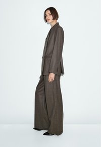 Brauner, maßgeschneiderter Anzug mit einem doppelt geknöpften Blazer und weit geschnittenen Hosen. Der Stoff ist strukturiert und das Design verfügt über auffällige Knöpfe.