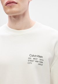 Homme portant un sweat-shirt blanc avec le texte Calvin Klein et l'adresse de New York imprimés sur la poitrine.