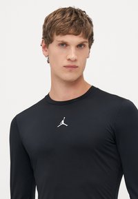 Jonge man met kort bruin haar die een aansluitend zwarte longsleeve shirt draagt met een klein wit logo van een basketballer op de borst.
