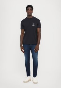 Tricou negru din bumbac cu un logo alb pe piept, asortat cu blugi albaștri strâmți și adidași de culoare crem. Fundal simplu.