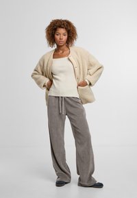 Fluffy crèmekleurige cardigan met grote zakken, gedragen over een aansluitend crèmekleurig ribbelshirt en een grijze wijde broek. Platte donkerblauwe schoenen maken de outfit compleet.