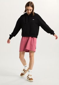 Svart oversized hoodie med en vit logotyp, parat med en knälång rosa kjol. Bärs med vita strumpor och bruna sneakers.