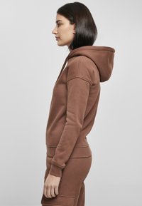Brun hettegenser laget av bomullsliknende materiale, med en frontlomme, ribbede mansjetter og hette. Designet er minimalistisk og figursydde.