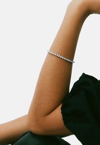 Rhinestone apyrankė ant riešo, sidabro tono metalas, su tolygiai išdėstytais apvaliais akmenimis. Po apyranke matoma odos spalva.