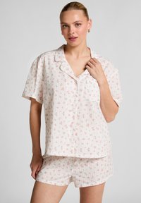 Vrouw in een witte korte mouwen knopen-blouse en bijpassende shorts met een klein roze bloemenpatroon, poserend met haar hand bij de kraag.