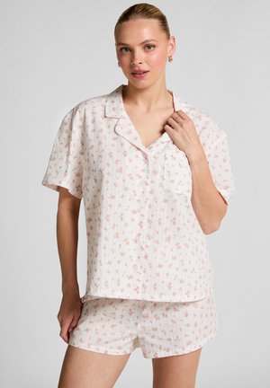 Vrouw in een witte korte mouwen knopen-blouse en bijpassende shorts met een klein roze bloemenpatroon, poserend met haar hand bij de kraag.