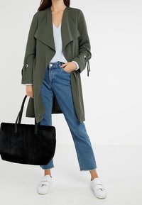 Olivově zelený dlouhý kabát s drapovaným límcem a nastavitelnými rukávy, v kombinaci s světle modrým texturovaným topem, denimovými džíny a černou taškou.