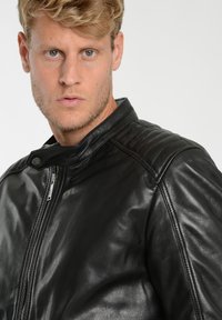 Sort læder motorcykeljakke med stående krave, kviltede skulderdetailler og sølv lynlås lukning. Glat, blank overflade.