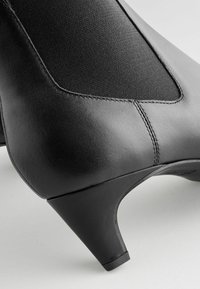 Gros plan sur une bottine en cuir noir avec un talon bas et effilé, ainsi qu'un panneau en tissu élastique pour un ajustement parfait.