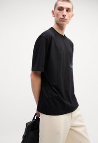 Tricou negru din bumbac, cu mâneci scurte și o buzunar pe piept, având literi brodate de culoare albastră. Modelul poartă pantaloni bej și are o geantă neagră.