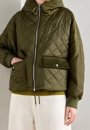 Persoon die een olijfgroene doorgestikte jas draagt met grote voorzakken en een ritssluiting aan de voorkant, gecombineerd met lichtgekleurde broek.