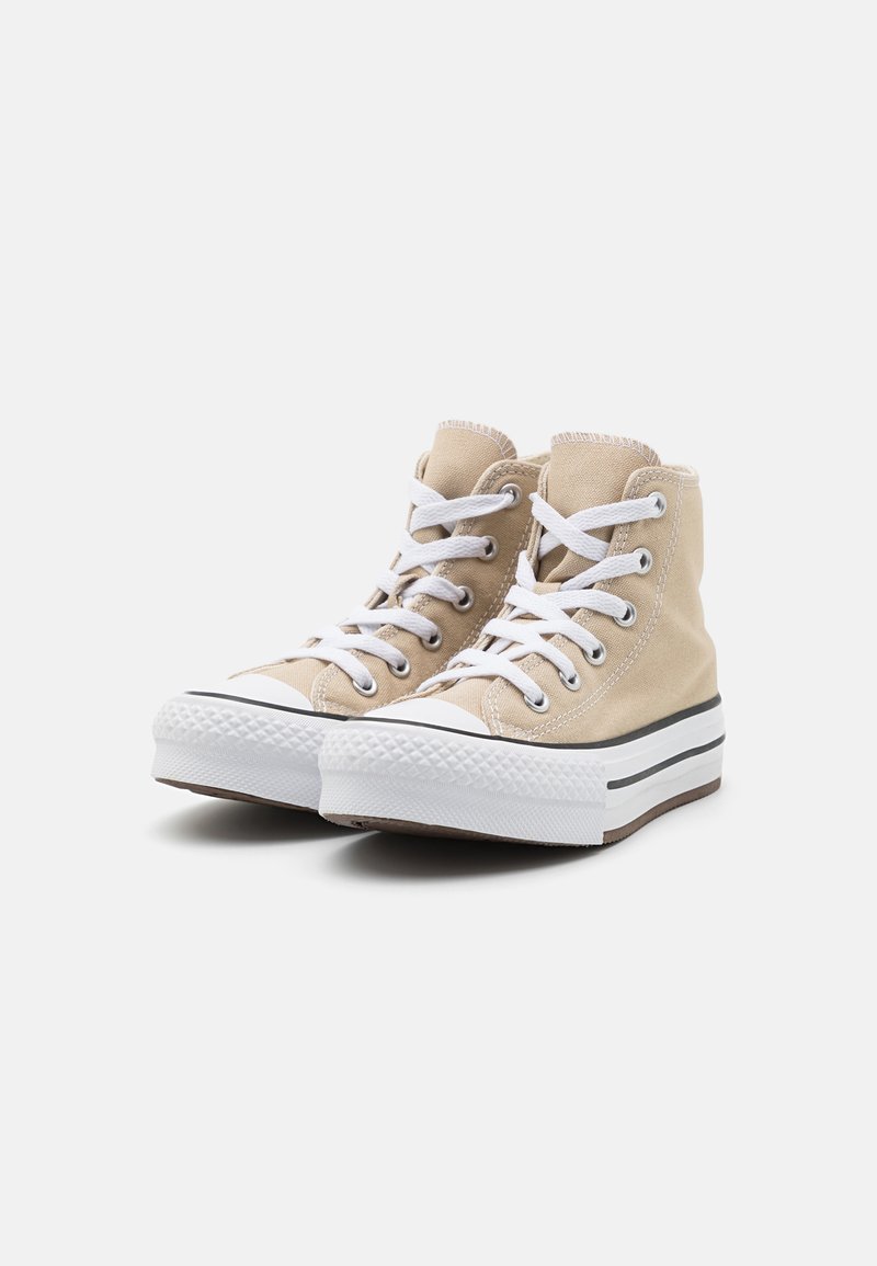 Zapatillas altas beige con cordones blancos, punteras de goma y parte superior de lona texturizada. Suela a rayas blancas y negras.