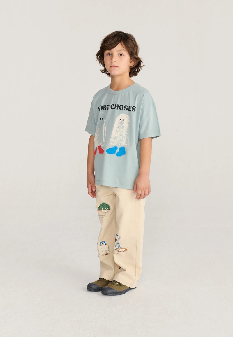 Enfant debout portant un t-shirt graphique bleu avec deux dessins de fantômes, un pantalon beige avec des patchs imprimés et des chaussures vertes.