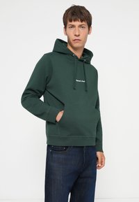 Felpa verde scuro con tasca a marsupio, cappuccio con cordoncino e logo ricamato. Abbinata a jeans in denim scuro. Tessuto morbido.