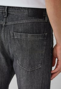 Graue Jeans aus Denim besitzen eine Gesäßtasche und ein schwarzes Lederlabel am Bund. Der Stoff hat eine dezente Textur und ein verblasstes Muster.