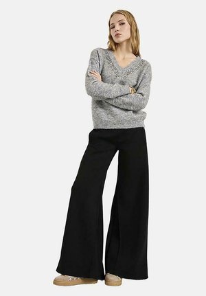Una mujer está de pie con los brazos cruzados, lleva un jersey gris con escote en V, pantalones negros de pierna ancha y zapatos beige sobre un fondo liso.