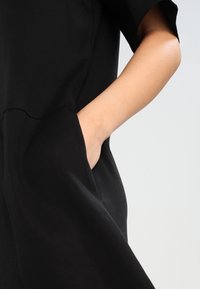 Robe noire à coupe ample, manches courtes et poches sur les côtés. Texture lisse, design simple et silhouette décontractée.