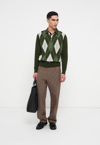 Grüner Argyle-Pullover mit weißen und hellgrünen Rauten, kombiniert mit braunen gestreiften Hosen und schwarzen Schuhen, dazu eine schwarze Tasche.