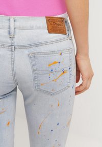 Vaqueros de mezclilla azul claro con manchas de pintura naranja y azul, que presentan una etiqueta de cuero marrón de Polo Ralph Lauren en la zona del bolsillo trasero.