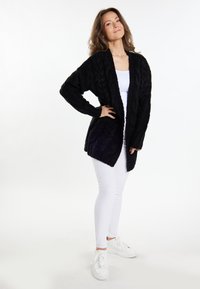 Cardigan nero in maglia con un motivo a treccia testurizzato, davanti aperto e maniche lunghe, abbinato a pantaloni bianchi e sneakers bianche.