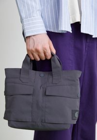 Harmaa kangaskassi, jossa on kaksi lyhyttä kahvaa, etutaskun läpät ja logopatch. Sileä pinta, jossa näkyvät ompeluyksityiskohdat.