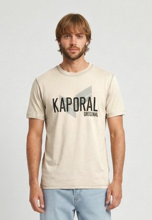 Homme aux cheveux châtain clair et barbe portant un t-shirt beige Kaporal Original et un jean bleu clair sur un fond gris uni.