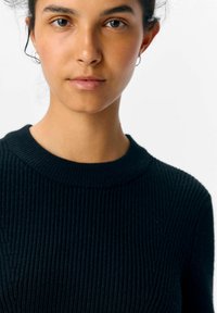 Schwarzer gerippter Strickpullover mit Rundhalsausschnitt, der eine strukturierte Oberfläche aufweist. Das Model hat natürliche Haare und minimalen Schmuck.