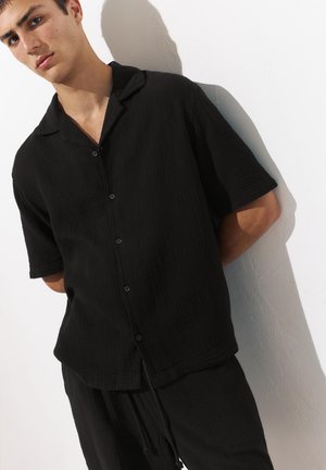 Jeune homme appuyé contre un mur blanc portant une chemise noire ample à manches courtes boutonnée et un pantalon noir assorti.